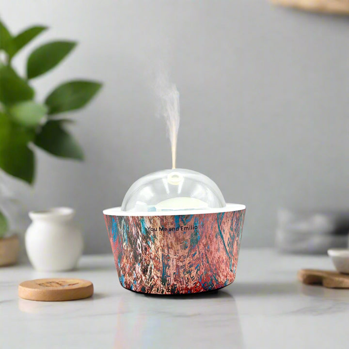 Artistic Essence Oil Diffuser, a masterful creation from Emilio Horton, founder of YOU ME & EMILIO. Featuring a vibrant, textured abstract design of deep blues, fiery reds, and shimmering gold, this diffuser transforms any space into a sanctuary of creativity, relaxation, and sensory delight.

More than just an aromatherapy tool, the Artistic Essence brings an immersive experience with the gentle sound of running water and soft mist creation, inspired by the tranquility of natural streams.