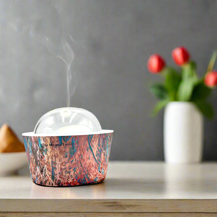 Artistic Essence Oil Diffuser, a masterful creation from Emilio Horton, founder of YOU ME &amp; EMILIO. Featuring a vibrant, textured abstract design of deep blues, fiery reds, and shimmering gold, this diffuser transforms any space into a sanctuary of creativity, relaxation, and sensory delight.

More than just an aromatherapy tool, the Artistic Essence brings an immersive experience with the gentle sound of running water and soft mist creation, inspired by the tranquility of natural streams.