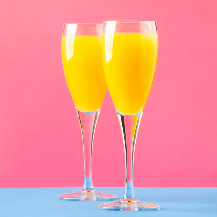 Two champagne flutes filled with orange juice on a pink and blue background