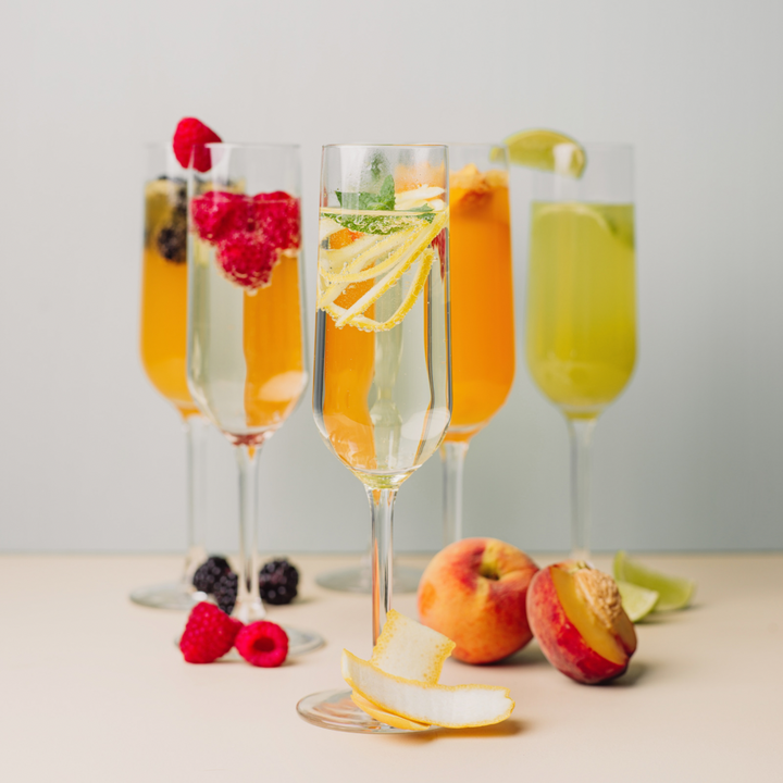 Five champagne flutes with various drinks and fruits on a light surface.