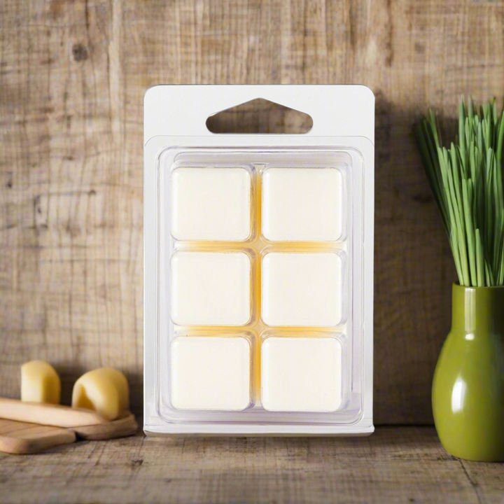 Pack of wax melts in a clear windowed packaging on a wooden surface with a vase and bread in the background.