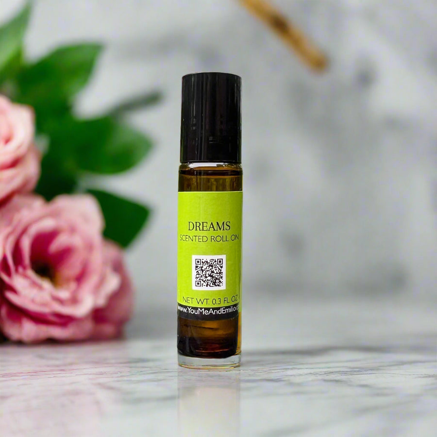 Amber glass bottle of Dreams Roll-On Perfume Oil by You Me & Emilio, featuring a blend of pink florals, soft blonde woods, and vanilla bean chiffon.